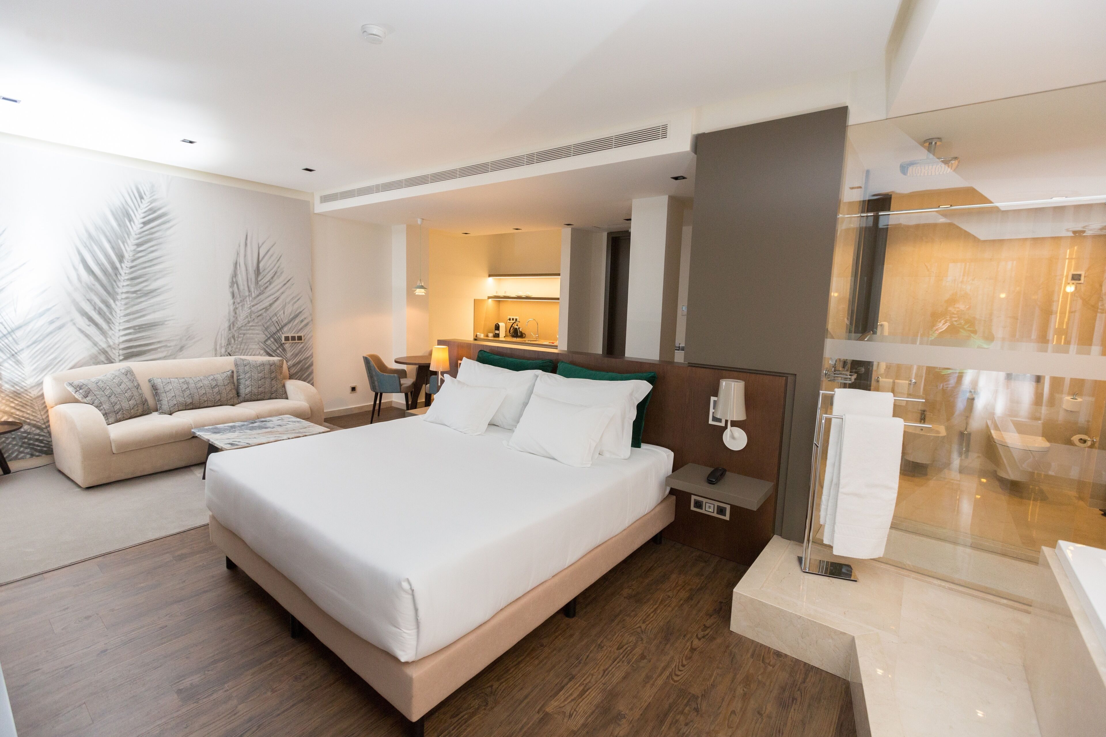 Una habitación de hotel moderna con una cama grande, un sofá, una mesita y un baño con paredes de vidrio.