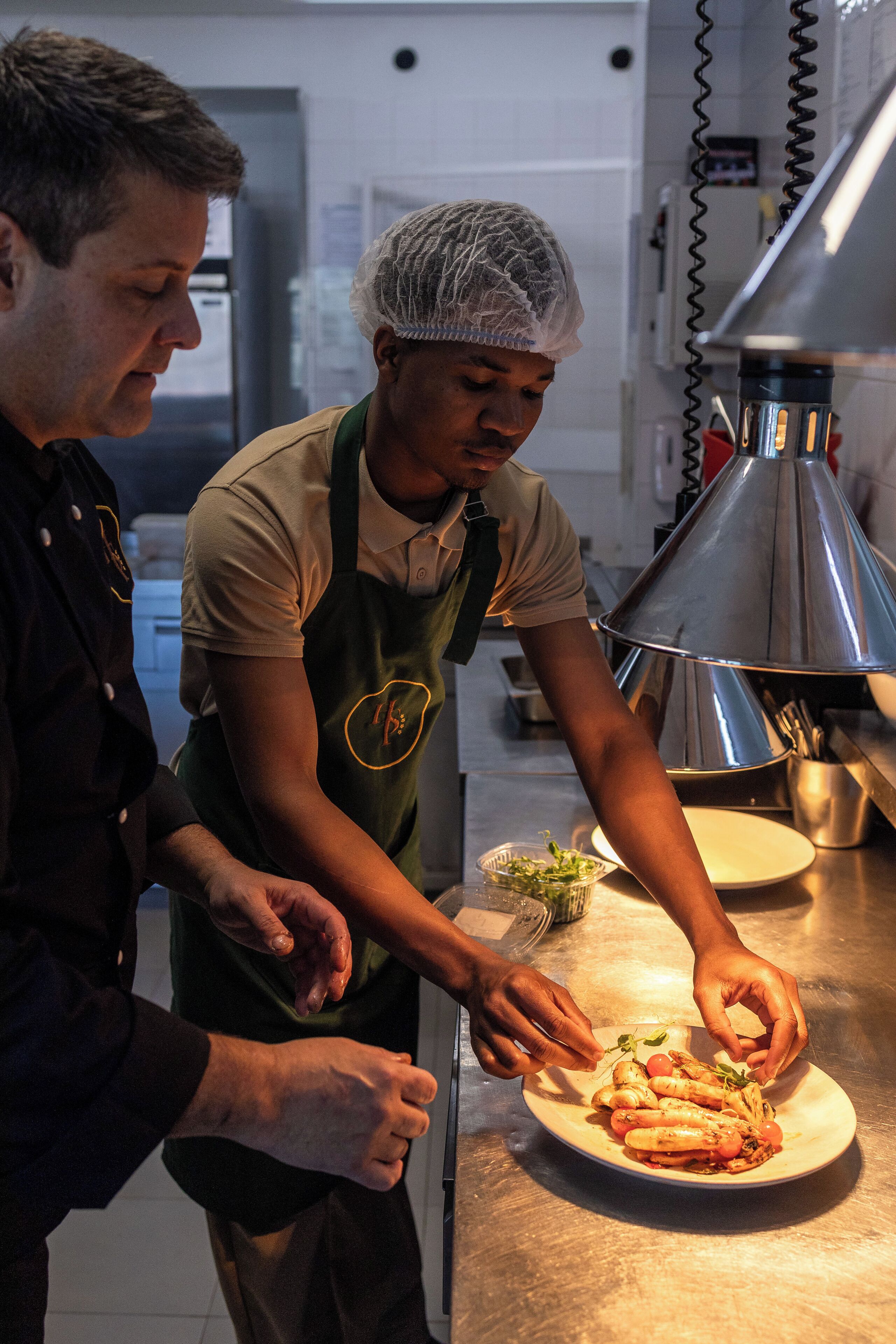 Two individuals in a kitchen preparing food on a stainless steel counter.