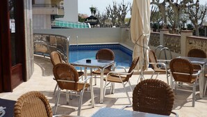 An outdoor pool area with wicker chairs and tables, a covered umbrella, and a view of the pool and surrounding trees.