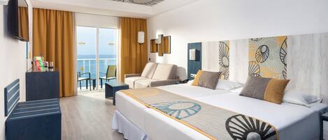 A hotel room with a large bed, a desk, a chair, and a balcony with a view of the sea.
