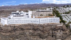Un grand hôtel moderne, doté de nombreux étages et de balcons, situé sur une falaise surplombant une zone côtière avec une plage et des eaux d’un bleu cristallin.