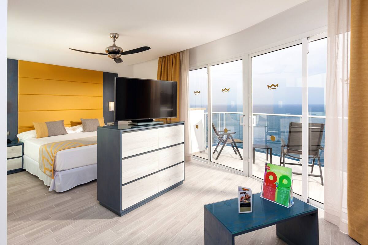 Ein Hotelzimmer mit Bett, Fernseher, Schreibtisch, einem Balkon mit Blick und einem Deckenventilator.