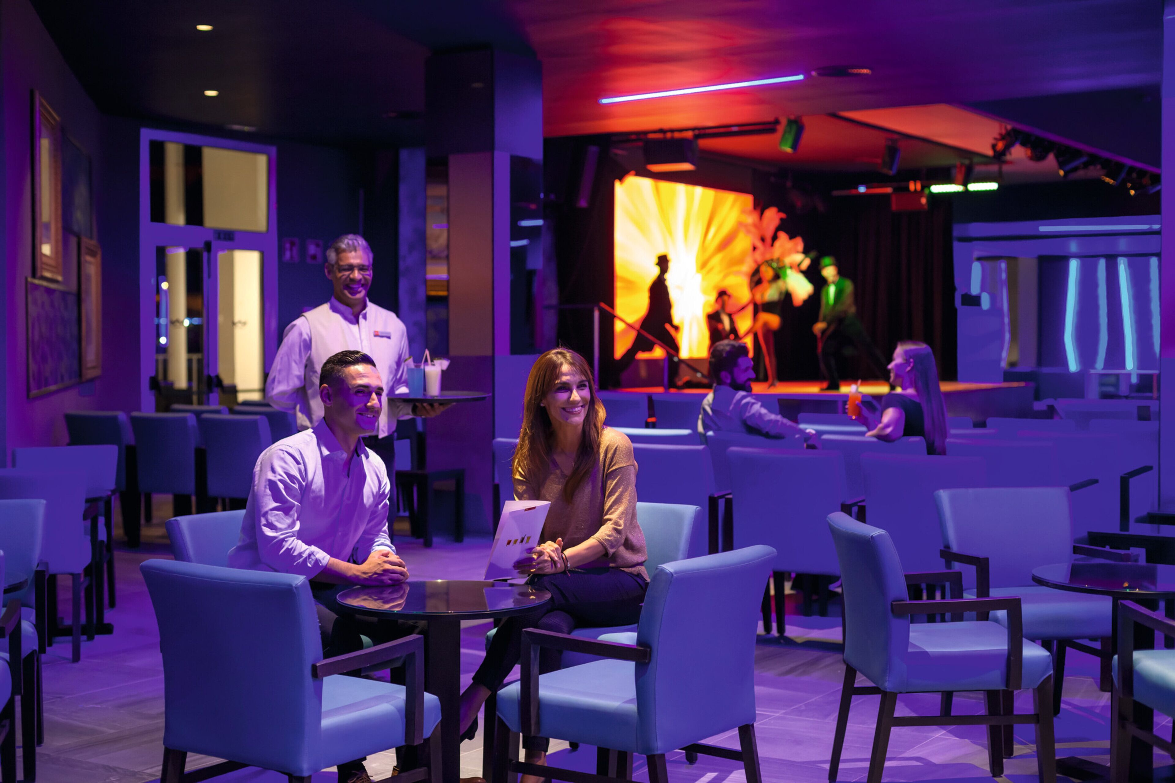 A modern lounge area with blue seating, a stage with performers, and a bar area.
