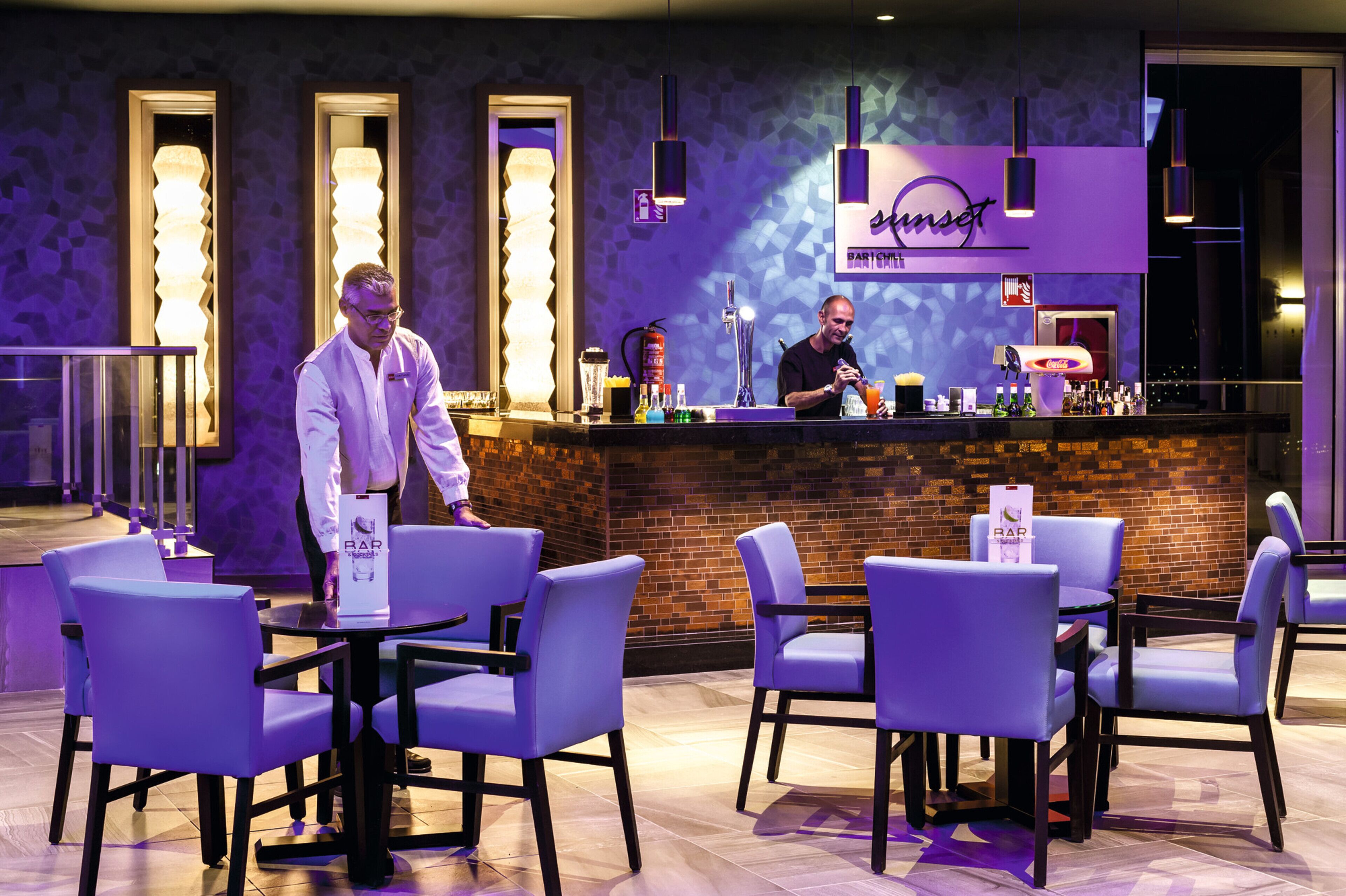 A bar area with a bartender, purple chairs, and a brick wall.