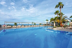 A swimming pool with lounge chairs and palm trees, overlooking a body of water. - Hotel Riu Vistamar - All Inclusive (Mogan)