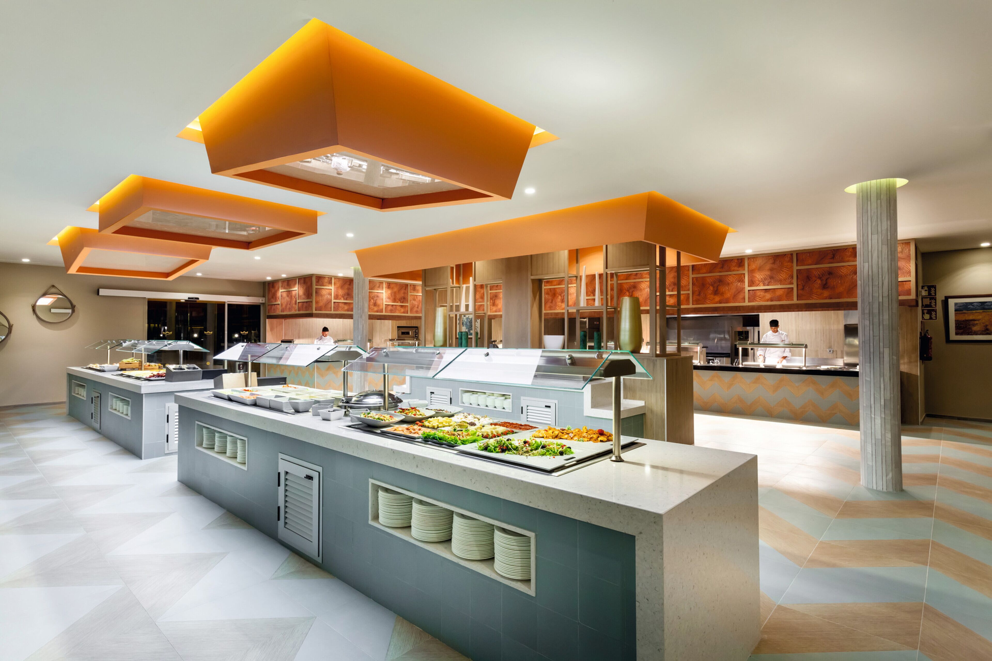 Zona de buffet moderna amb diverses estacions de servei, una illa central plena de menjar i un disseny de sostre contemporani.
