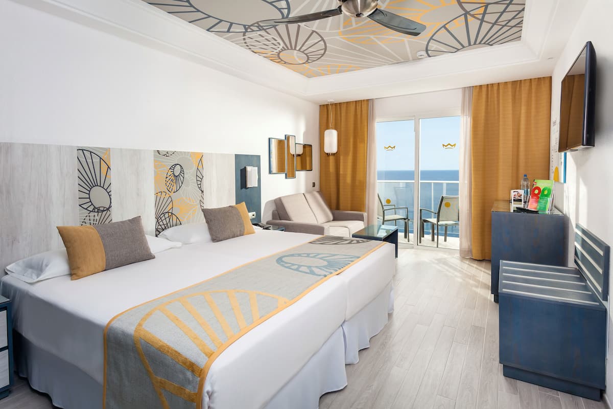 Ein Hotelzimmer mit einem großen Bett, einem Schreibtisch, einem Sessel, einem Fernseher und einem Balkon mit Meerblick.