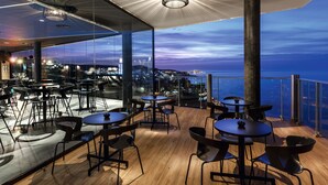 Un restaurant moderne offrant une vue sur la ville au crépuscule, avec des tables rondes et des chaises noires.