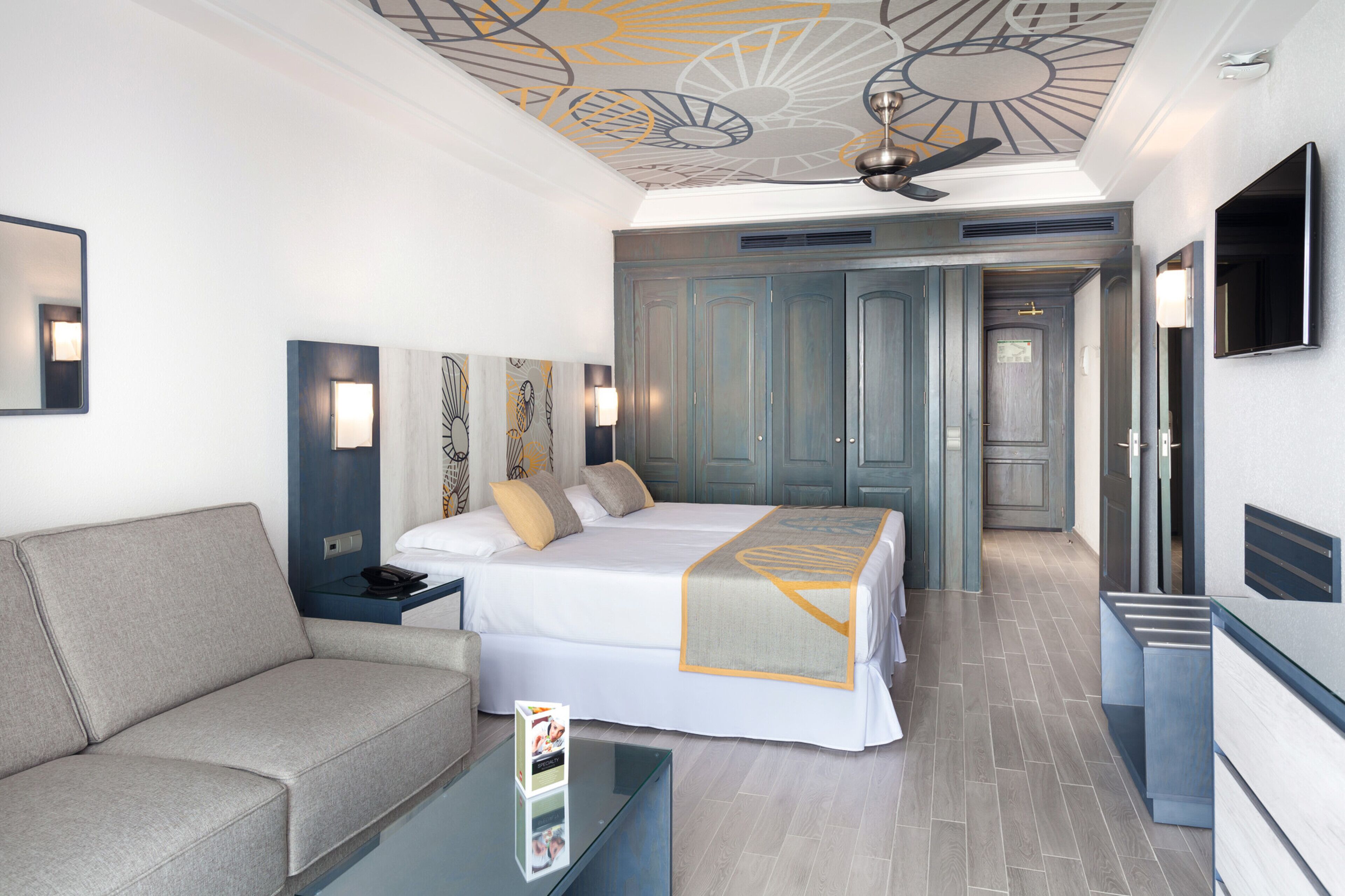 A modern hotel room with a large bed, a sofa, a glass coffee table, and a ceiling fan.
