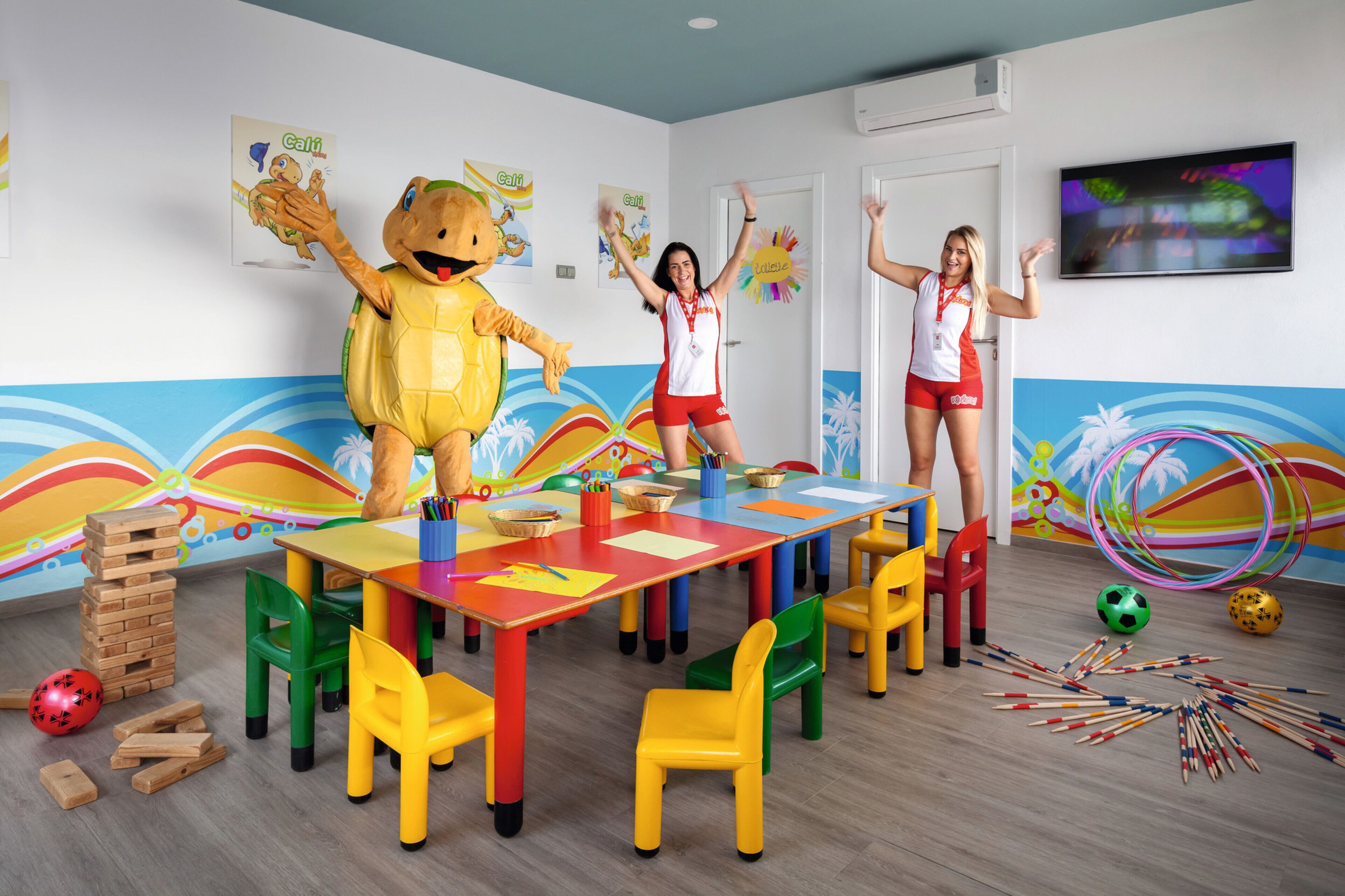 Un aula de colores brillantes con un personaje representativo, una mesa con material educativo y un televisor en la pared.