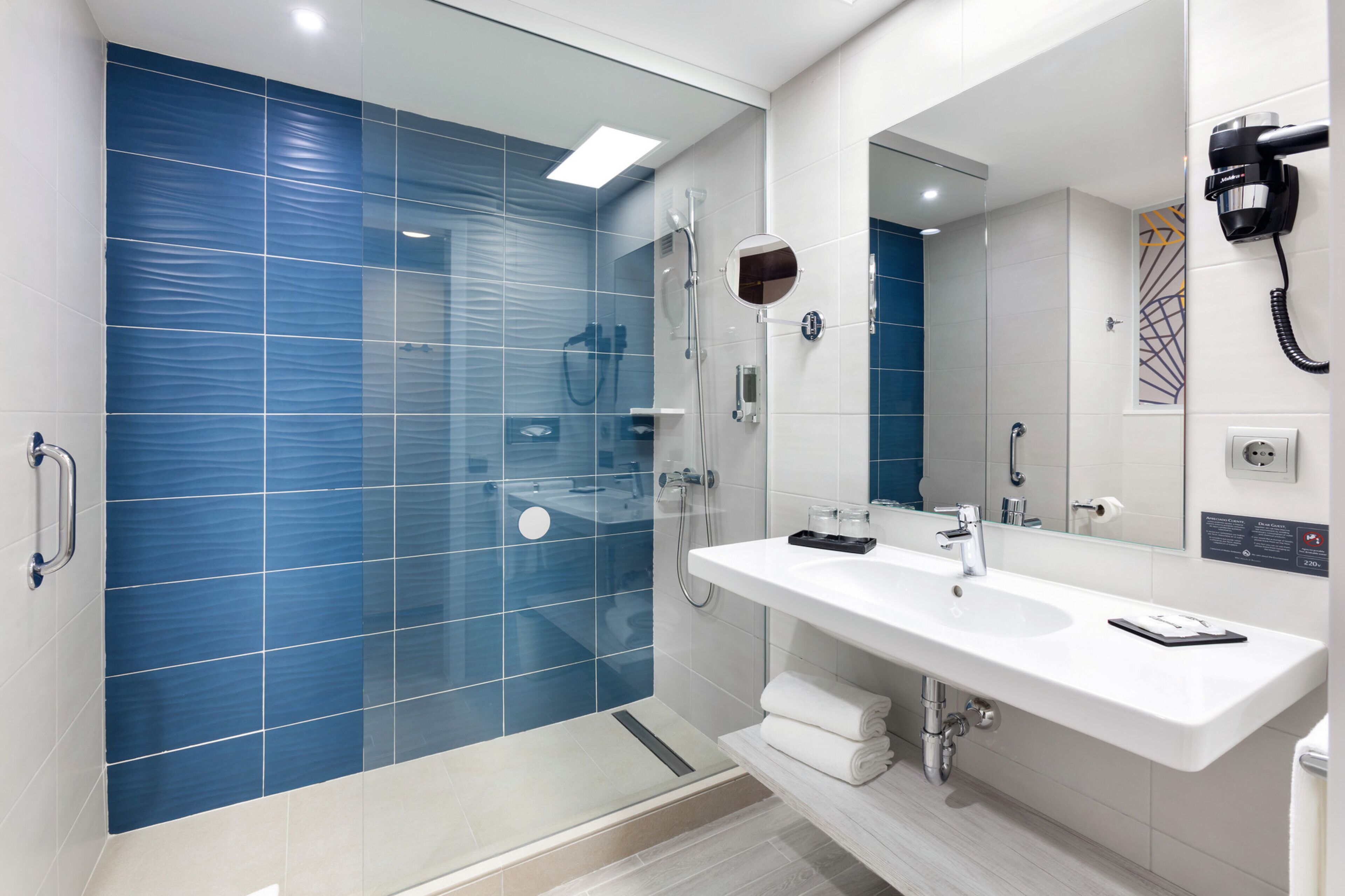 Un bagno moderno con cabina doccia in vetro, lavabo bianco con specchio e asciugacapelli montato a parete.