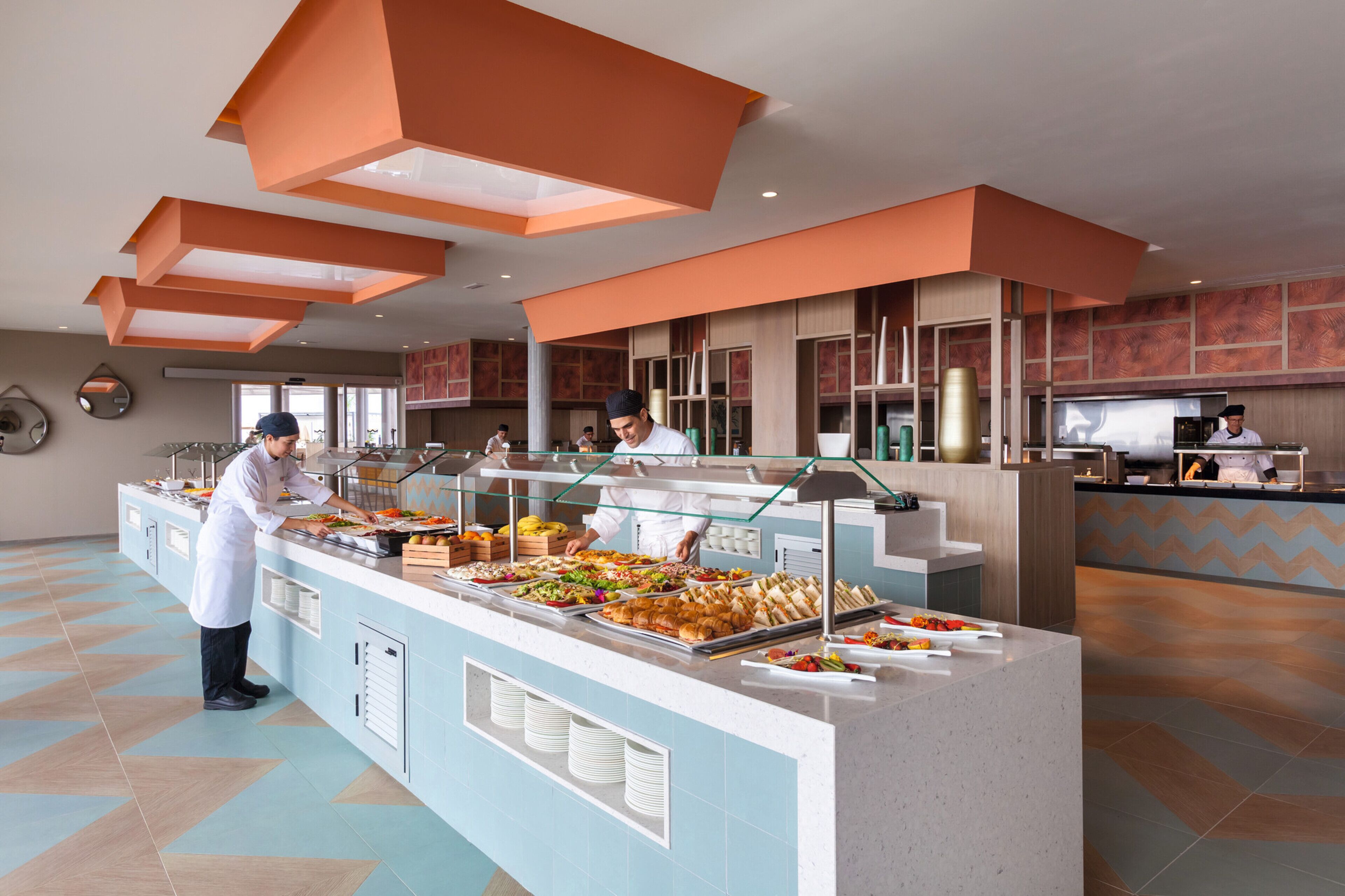 A buffet area with a variety of food items, a counter with a glass barrier, and a modern interior design with geometric ceiling structures.