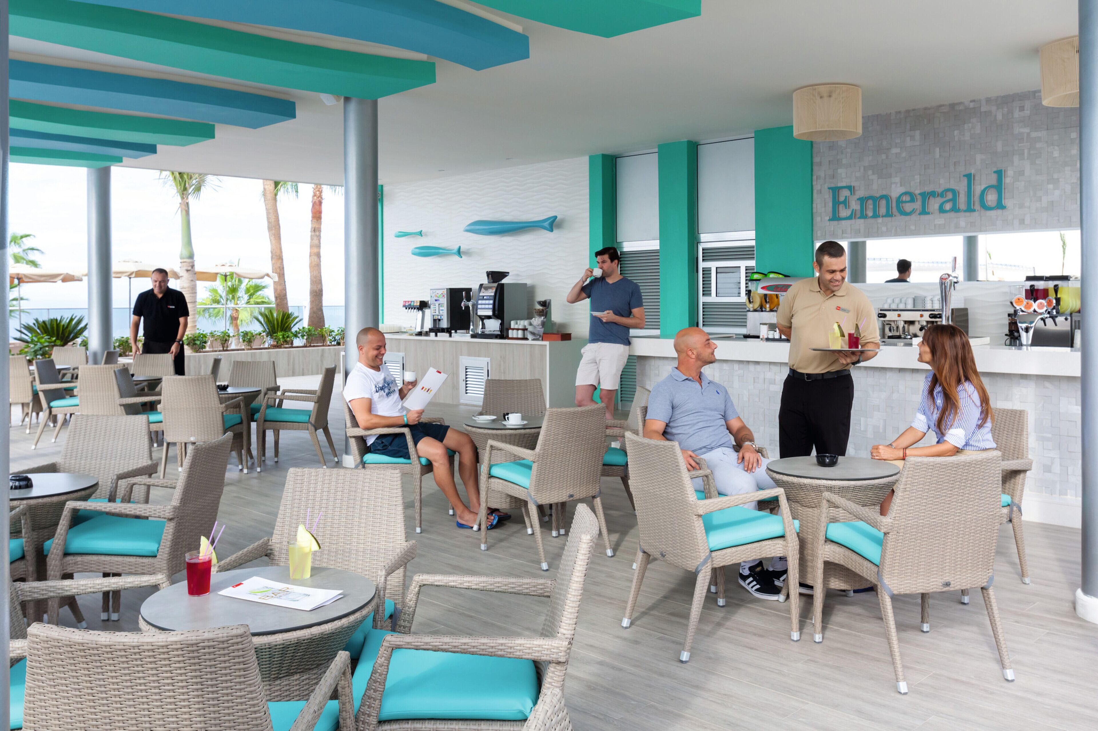 Un espace extérieur moderne, aménagé avec des meubles en osier et des coussins de couleur turquoise, un bar avec une enseigne bien visible, et une vue sur la mer.