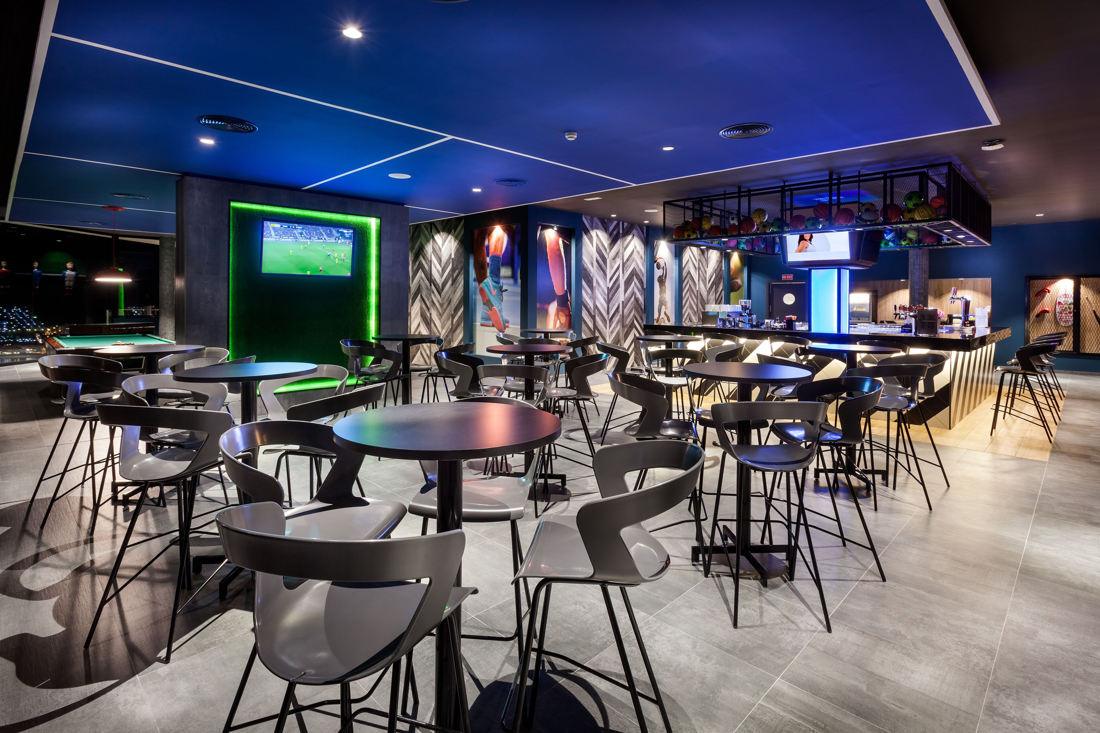 Un área de bar moderna con varias mesas y sillas, una pantalla grande mostrando un evento deportivo y un diseño de pared con un llamativo patrón de espiga.