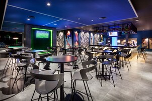 Un área de bar moderna con varias mesas y sillas, una pantalla grande mostrando un evento deportivo y un diseño de pared con un llamativo patrón de espiga.