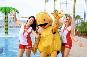 Un personaje publicitario con un disfraz amarillo y un atuendo rojo y blanco, de pie junto a una piscina con palmeras al fondo.