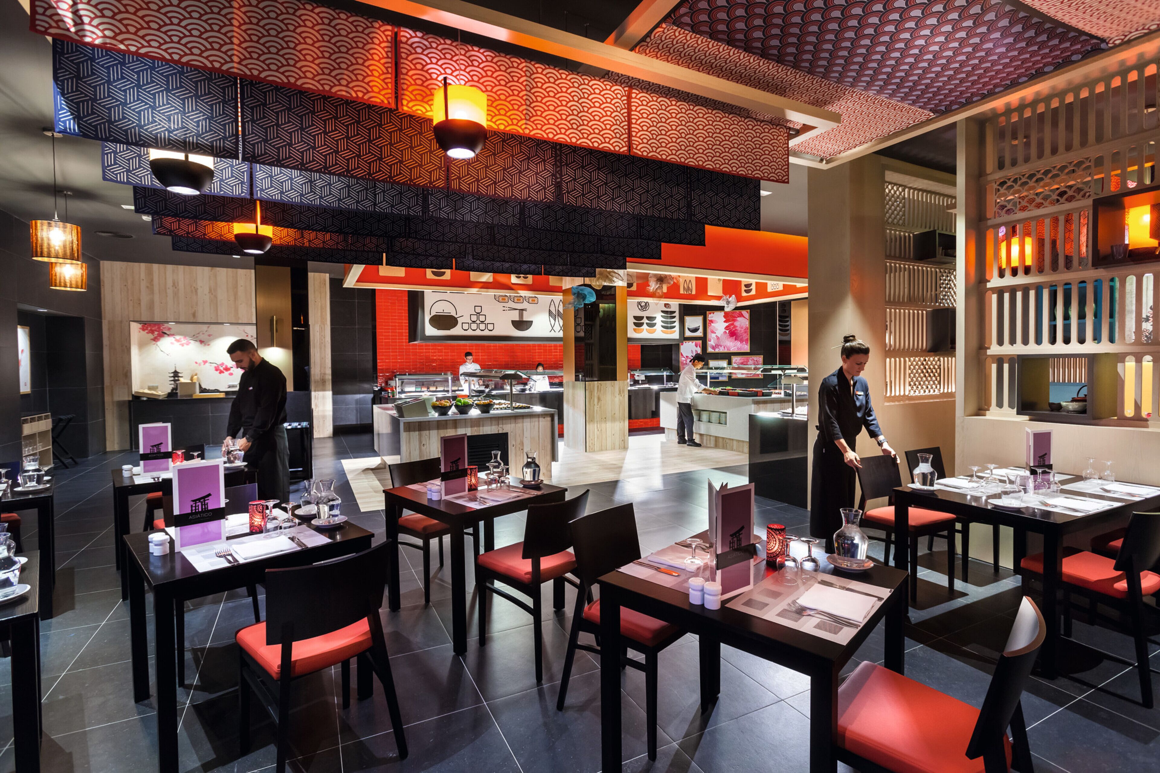 Un restaurante moderno con techo alto, diseños de pared estampados y una zona de cocina central. Las mesas están dispuestas con sillas rojas y manteles blancos.