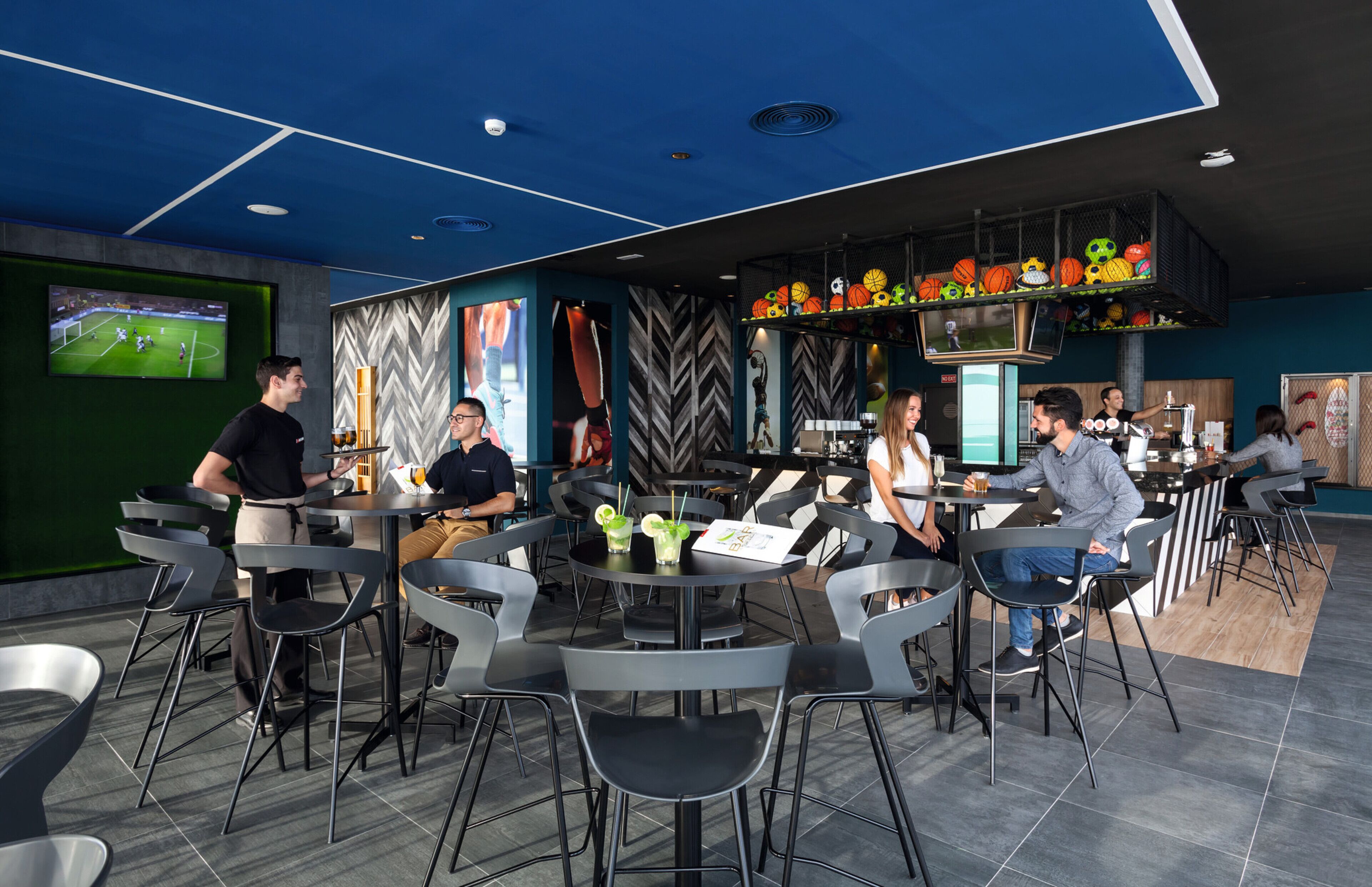 A modern restaurant with black chairs and tables, a large screen displaying a soccer game, and a variety of sports balls on display.