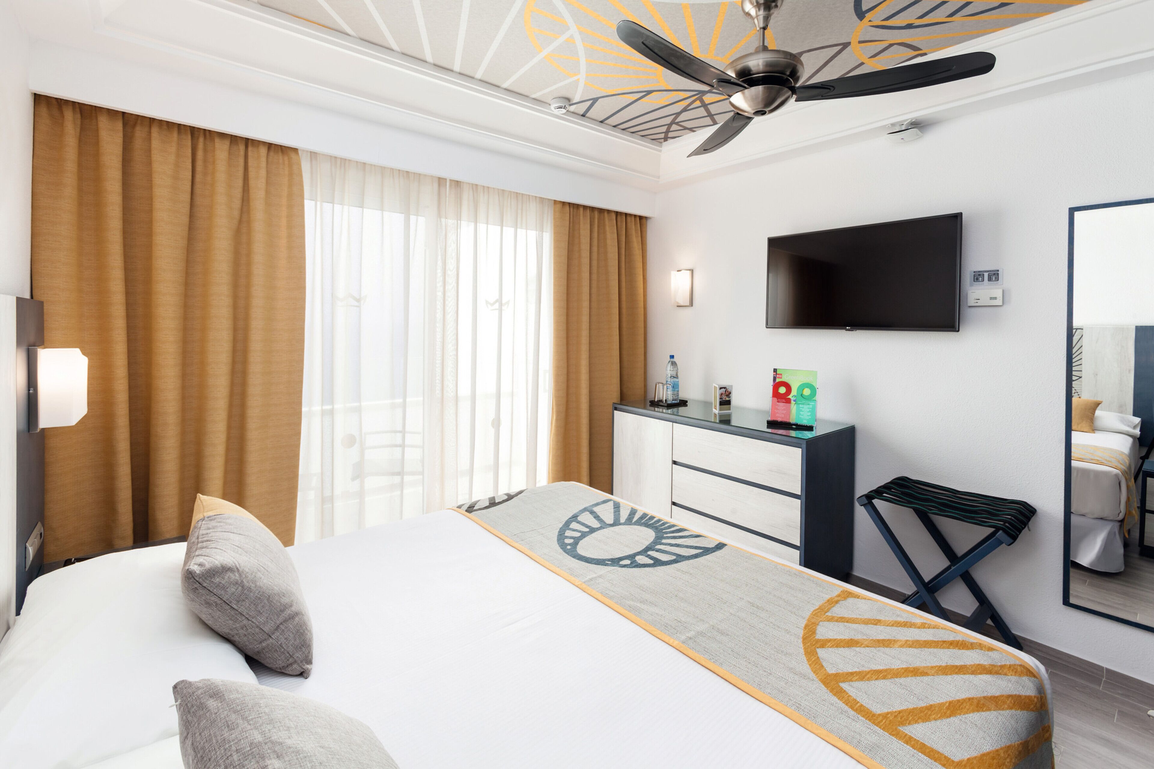 Een hotelkamer met een bed, bureau, flatscreen televisie, plafondventilator en een groot raam met gordijnen.