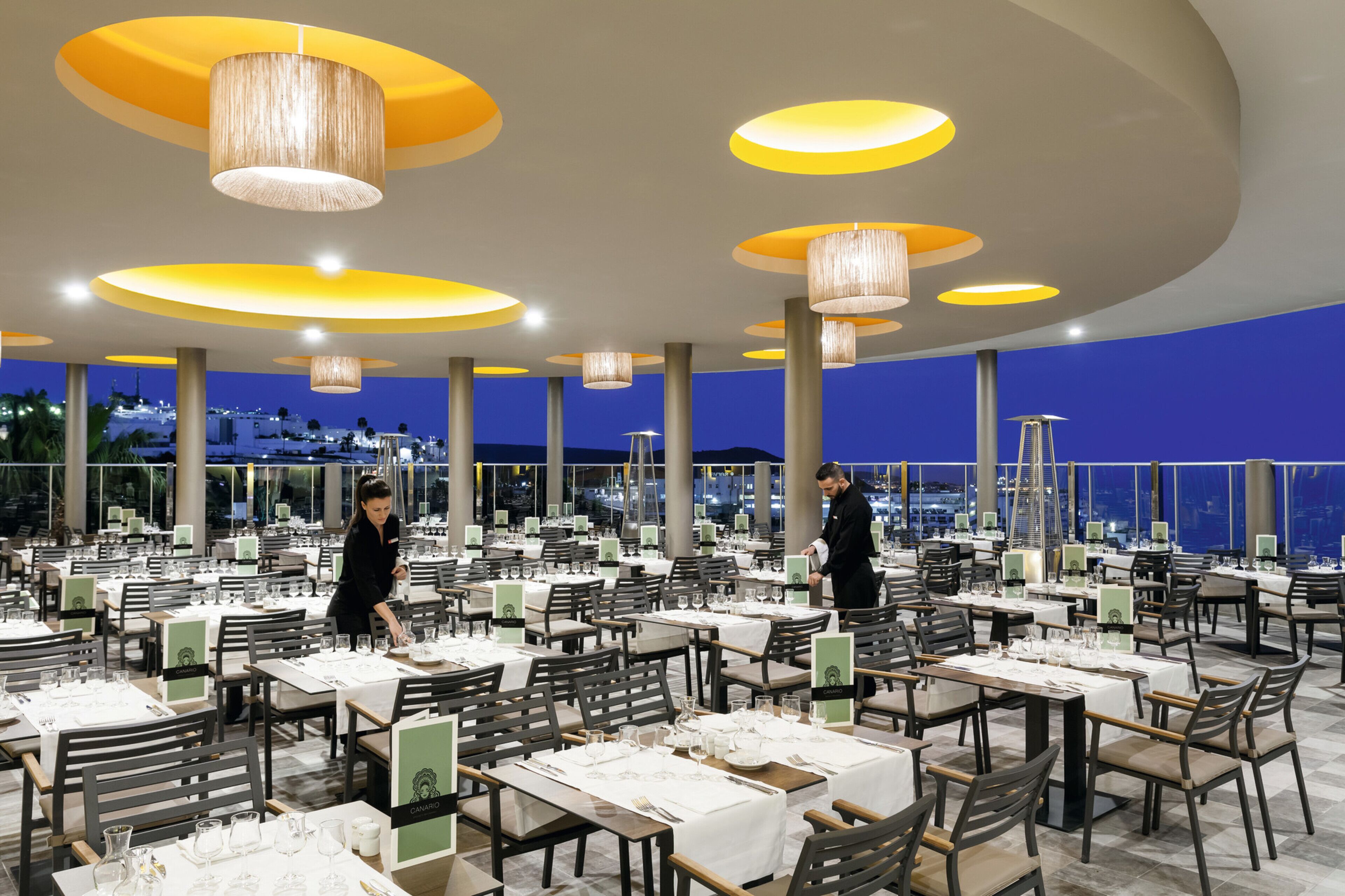 A spacious outdoor dining area with multiple tables and chairs, large hanging light fixtures, and a city view in the background.