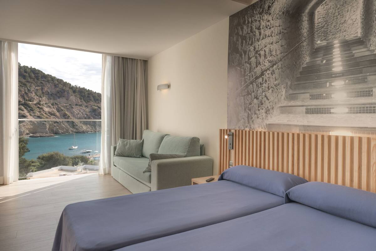 A modern hotel room with a large bed, a sofa, and a view of a coastal landscape.