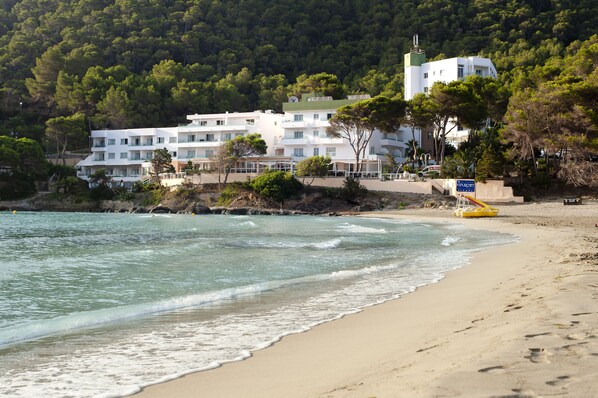 A coastal hotel with multiple white buildings, a sandy beach, and a forested hillside in the background. - Hotel Apartamentos El Pinar (Santa Eulalia del Rio)