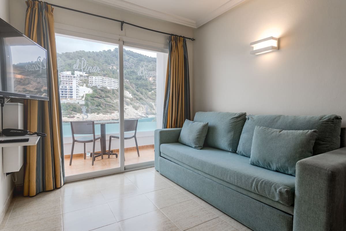 A modern living room with a sofa, a small table, and a chair. A balcony with a glass door leads to a terrace with a view of a beach and buildings.