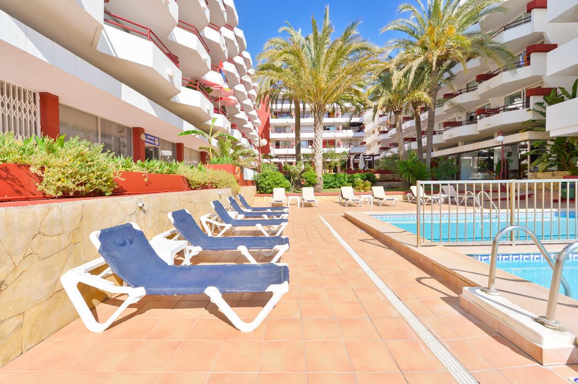 A poolside area with sun loungers, a swimming pool, and a building with multiple balconies and palm trees.