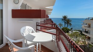 Varanda com cadeiras e mesa brancas, com vista para um mar azul e palmeiras.