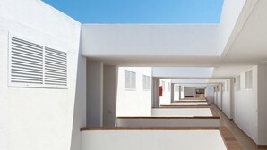 Um corredor comprido e estreito, com paredes brancas e piso de madeira, que dá acesso a vários quartos.