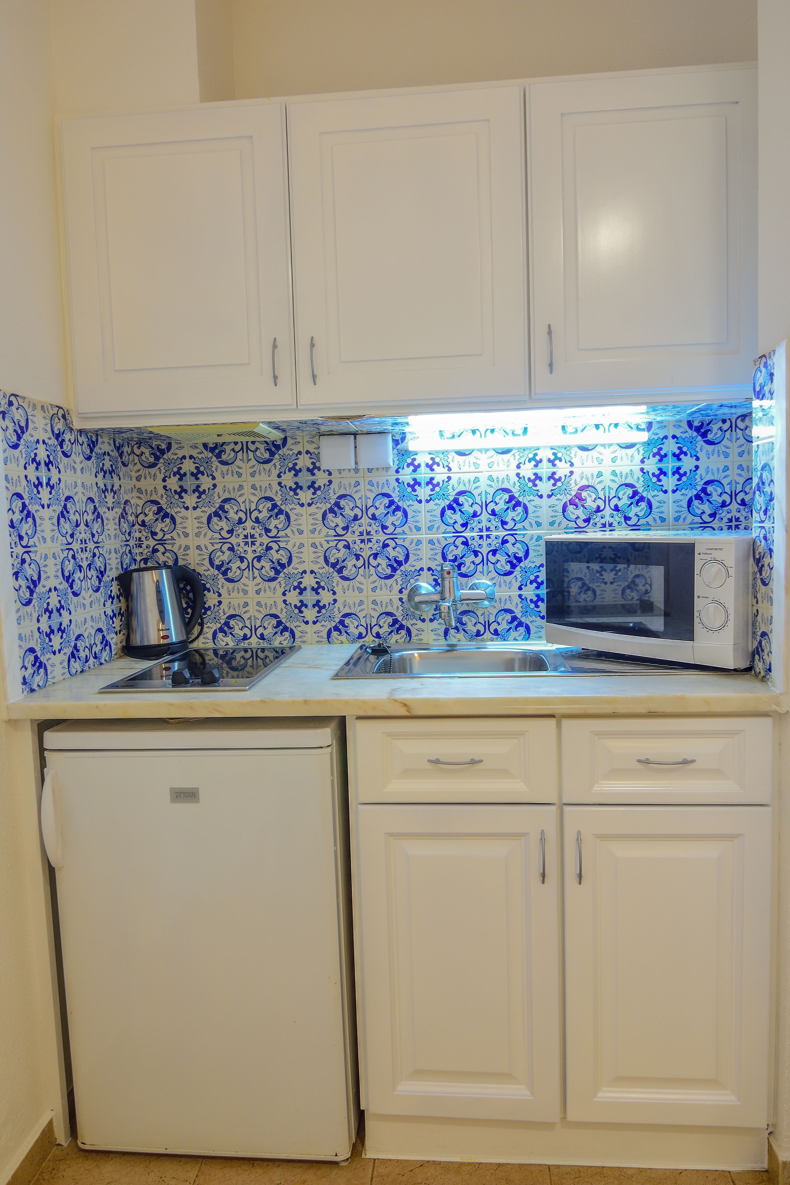 Una cocina compacta con armarios blancos, un microondas, un hervidor y un refrigerador pequeño.