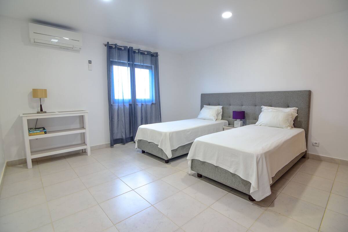 A hotel room with two beds, a bedside table, a shelf with books, and a window with curtains.