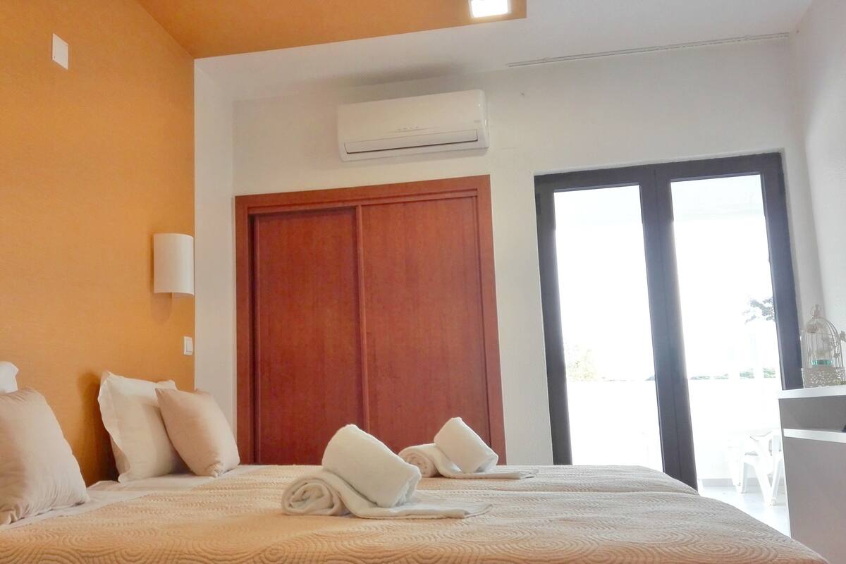 A hotel room with a bed, pillows, a wooden wardrobe, an air conditioning unit, and a view through a glass door.