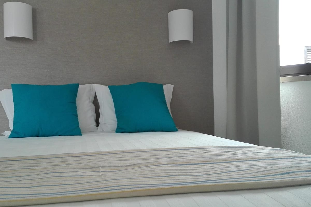 A modern hotel room with a bed, two teal pillows, a white bedspread, and two wall-mounted light fixtures.