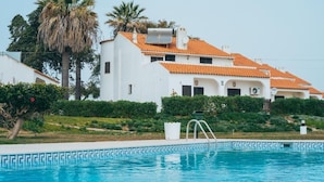 Un bâtiment blanc avec des toits recouverts de tuiles orange, une piscine aux carreaux de mosaïque bleus et des palmiers.