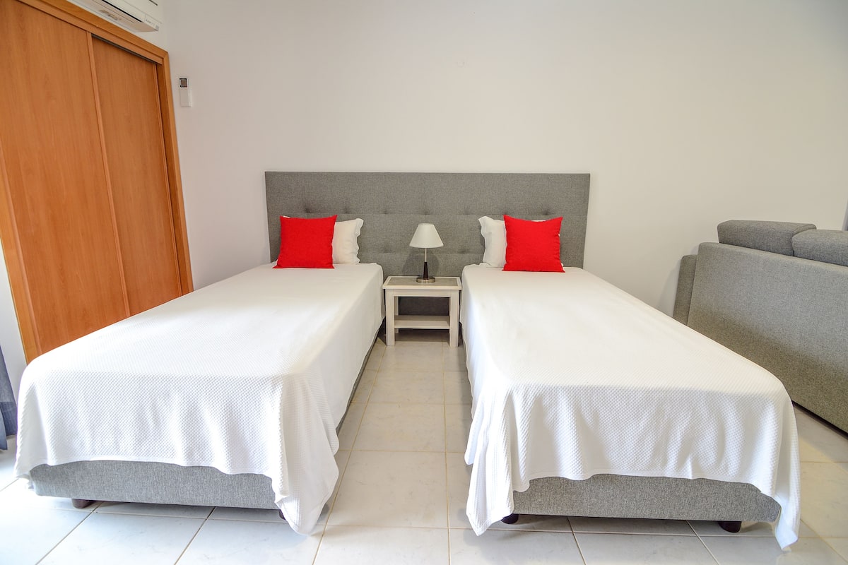 A double room with two beds, a nightstand, a lamp, and a grey sofa.