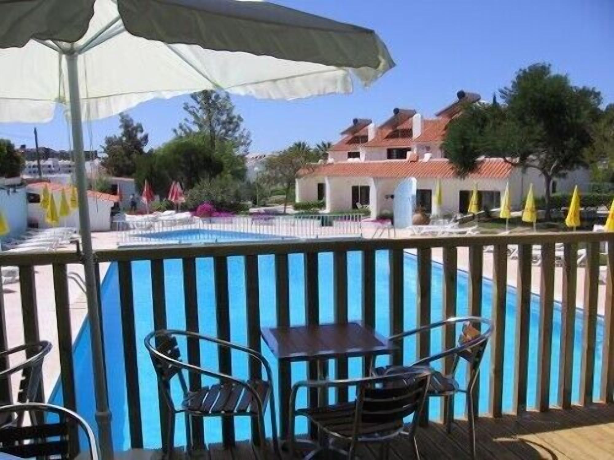 Un espace aménagé au bord de la piscine, avec une table et des chaises, un parasol blanc et une vue sur un bâtiment doté d’un toit rouge.