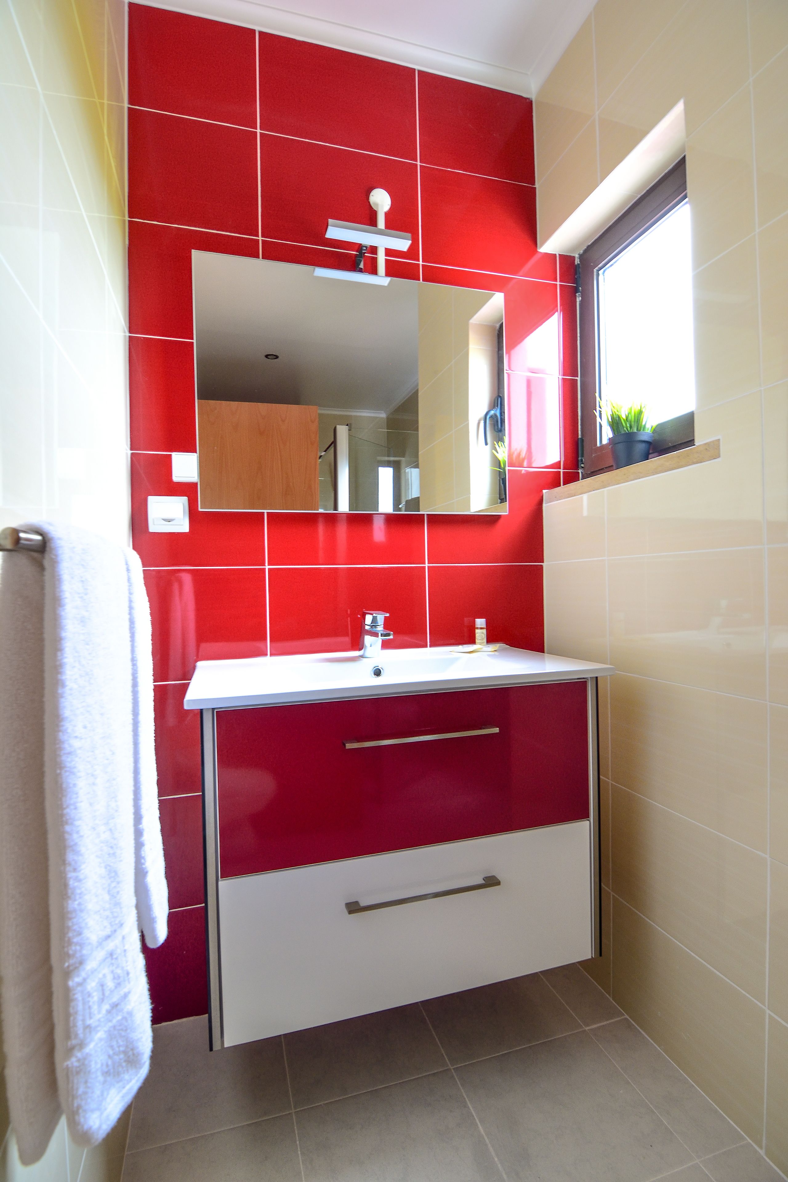Une salle de bain avec des carreaux rouges et beiges, un lavabo blanc, un miroir et une fenêtre avec une plante.