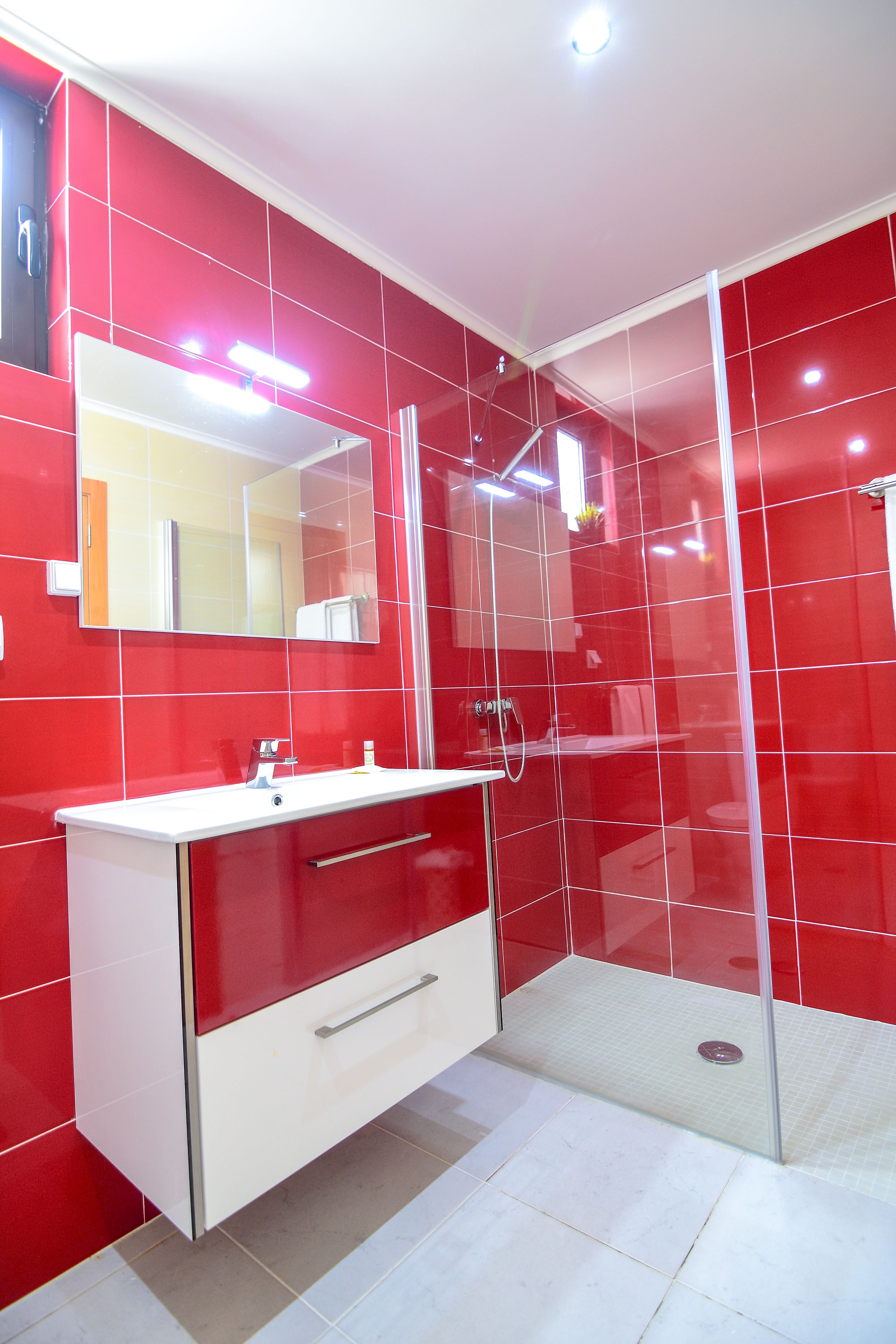 Ein modernes Badezimmer mit roten Fliesen, einem weißen Waschtisch, einer Glasdusche und einem Spiegel über dem Waschbecken.