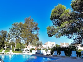 Una piscina con sillas de descanso, árboles y edificios al fondo.