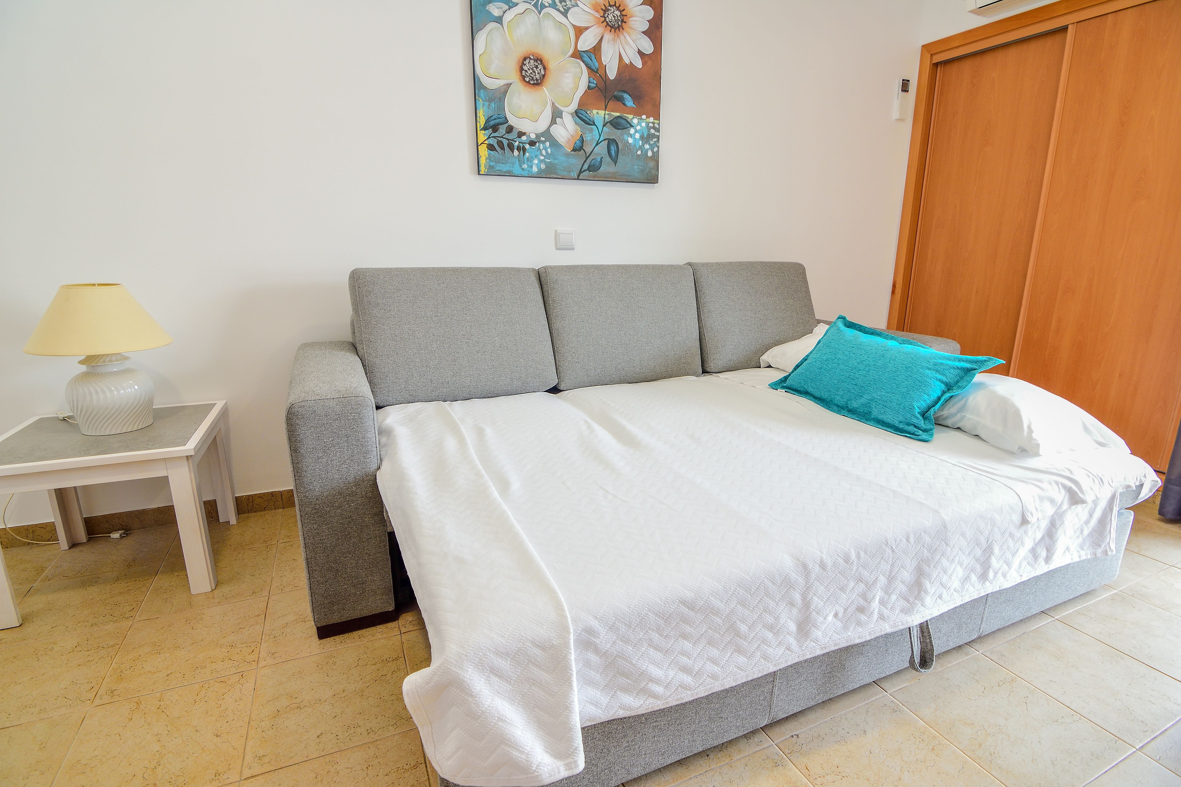 Une chambre à coucher avec un lit, un canapé gris, une petite table avec une lampe et une porte en bois.