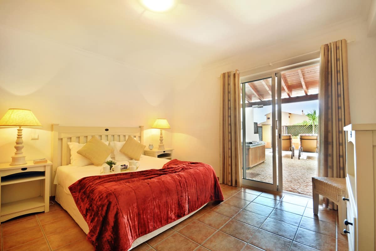 A hotel room with a bed, two bedside tables, a lamp, a red bedspread, and a sliding glass door leading to an outdoor area with chairs.