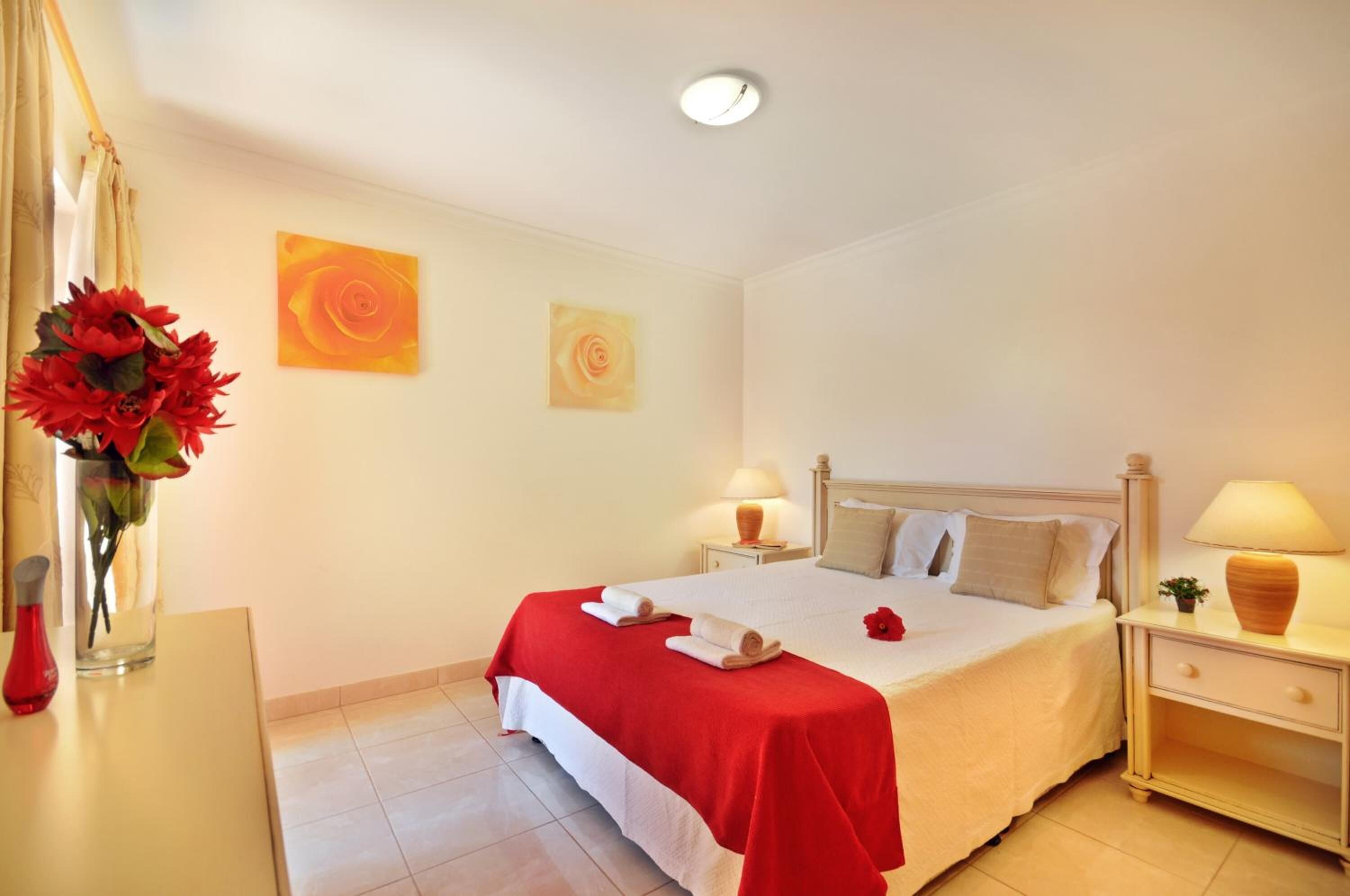 Quarto de hotel com uma cama, duas mesas de cabeceira, um criado-mudo, um vaso com flores e duas obras de arte nas paredes.