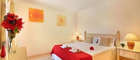 Quarto de hotel com uma cama, duas mesas de cabeceira, um criado-mudo, um vaso com flores e duas obras de arte nas paredes.