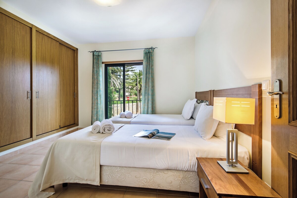 A hotel room with two beds, a wooden wardrobe, a bedside table with a lamp, and a view of the outdoors through a window with curtains.
