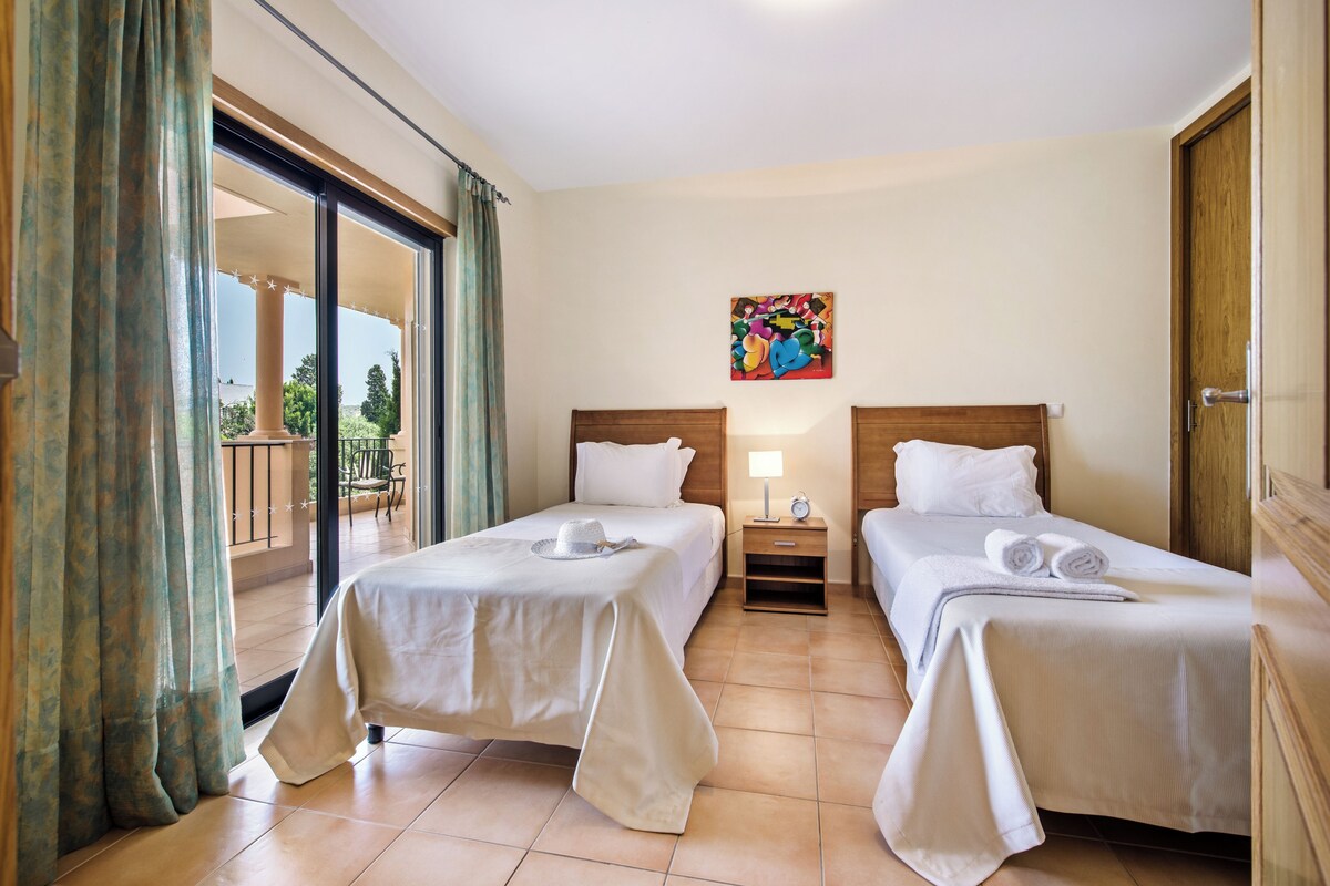 A hotel room with two beds, a balcony, a painting on the wall, and a nightstand with a lamp.