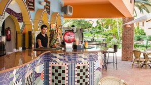 A bar area with a tiled counter, a Coca-Cola branded drink dispenser, and a view of a garden with palm trees and a brick wall. - Jardim Da Meia Praia Hotel (Lagos)