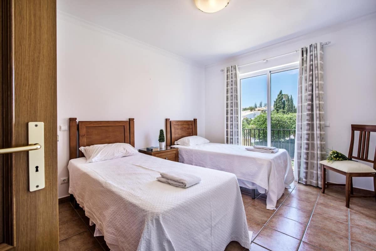 A hotel room with two beds, a balcony with curtains, and a view of greenery.