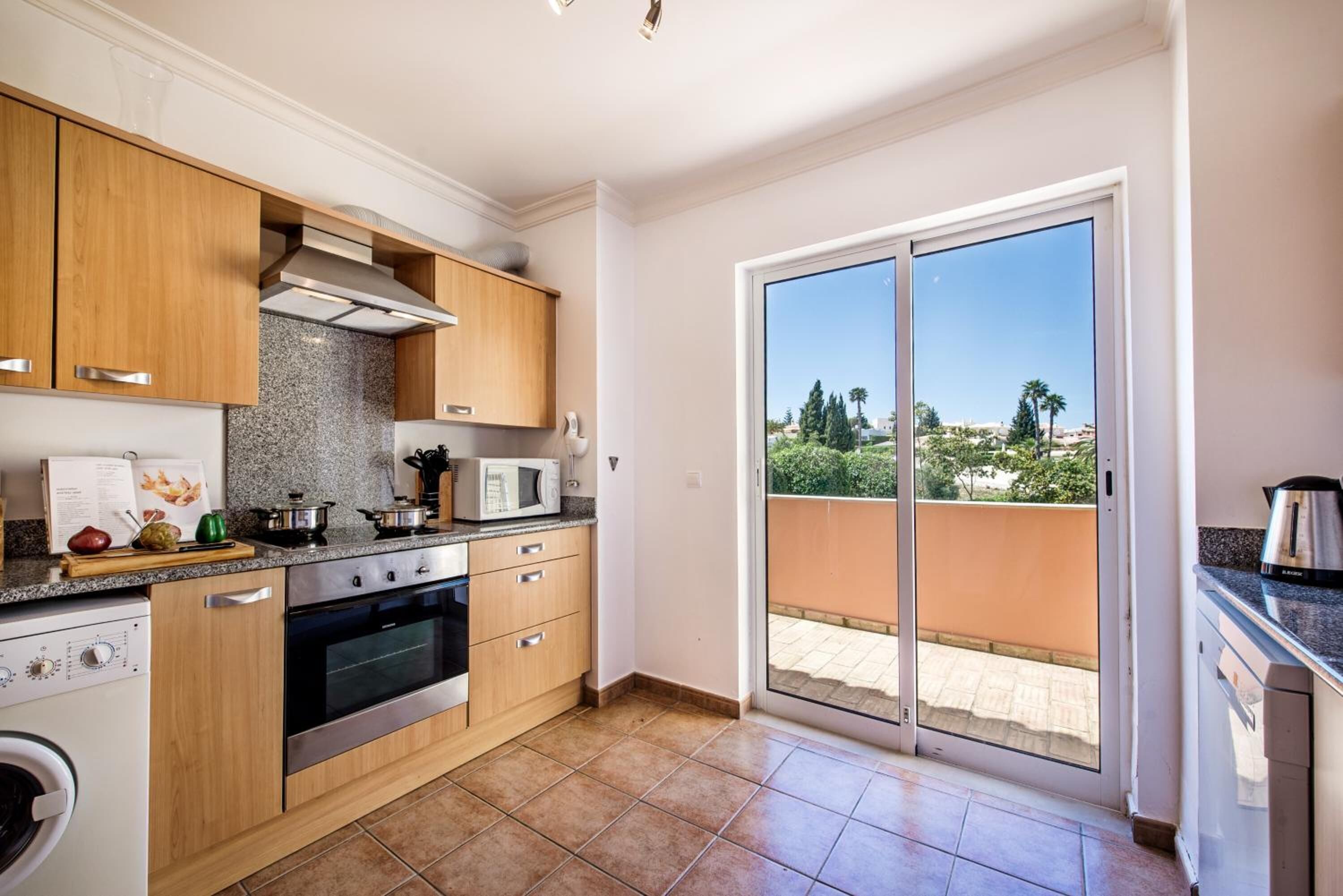 Eine moderne Küche mit Holzausstattung, einem Einbauofen und einer Mikrowelle. Ein großes Schiebeglastür führt zu einem Balkon mit Blick auf Bäume und Gebäude.