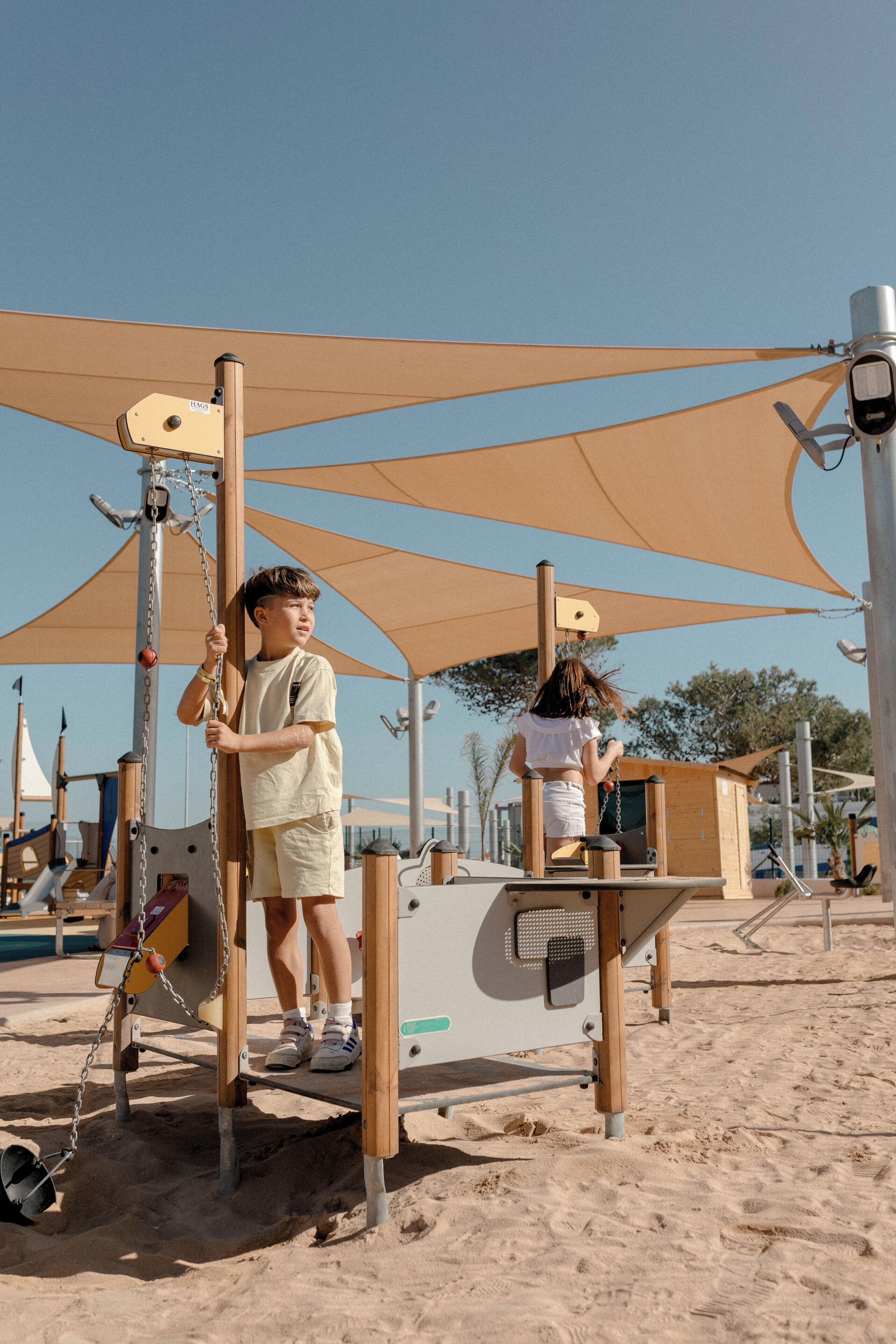 A playground with swings and climbing structures, surrounded by sand and shaded by large umbrellas.